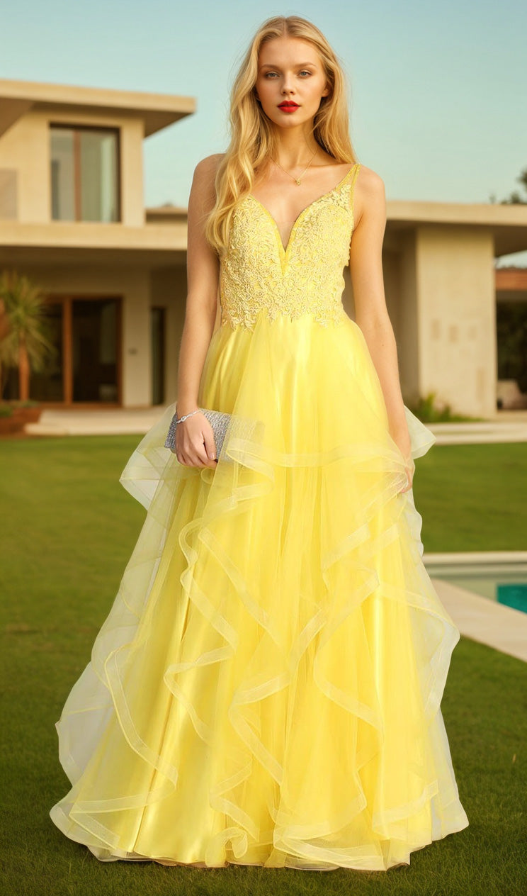 A woman stands on a lawn in front of a modern house, wearing the Sheer-Sides Long Tiered Prom Dress: Chicas XR6006, a yellow evening gown with a fitted bodice and tiered tulle skirt, holding a small clutch.